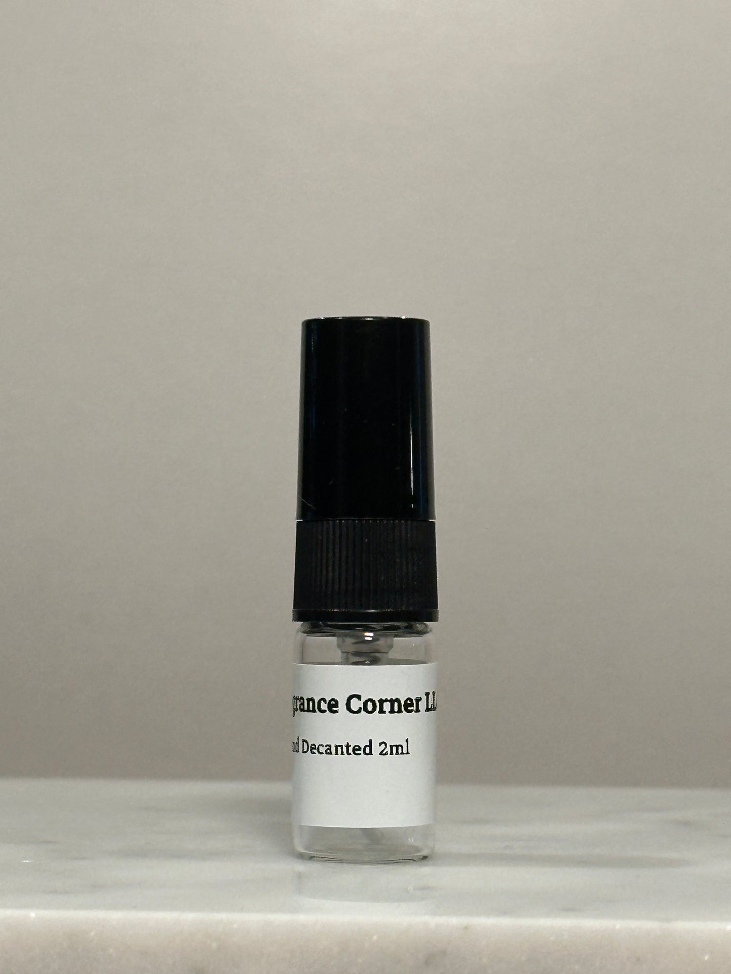 A small clear glass decanted perfume sample bottle with a black cap and a white label reading 'Prince Corner Lot Decanted 2ml', set against a neutral gray background.