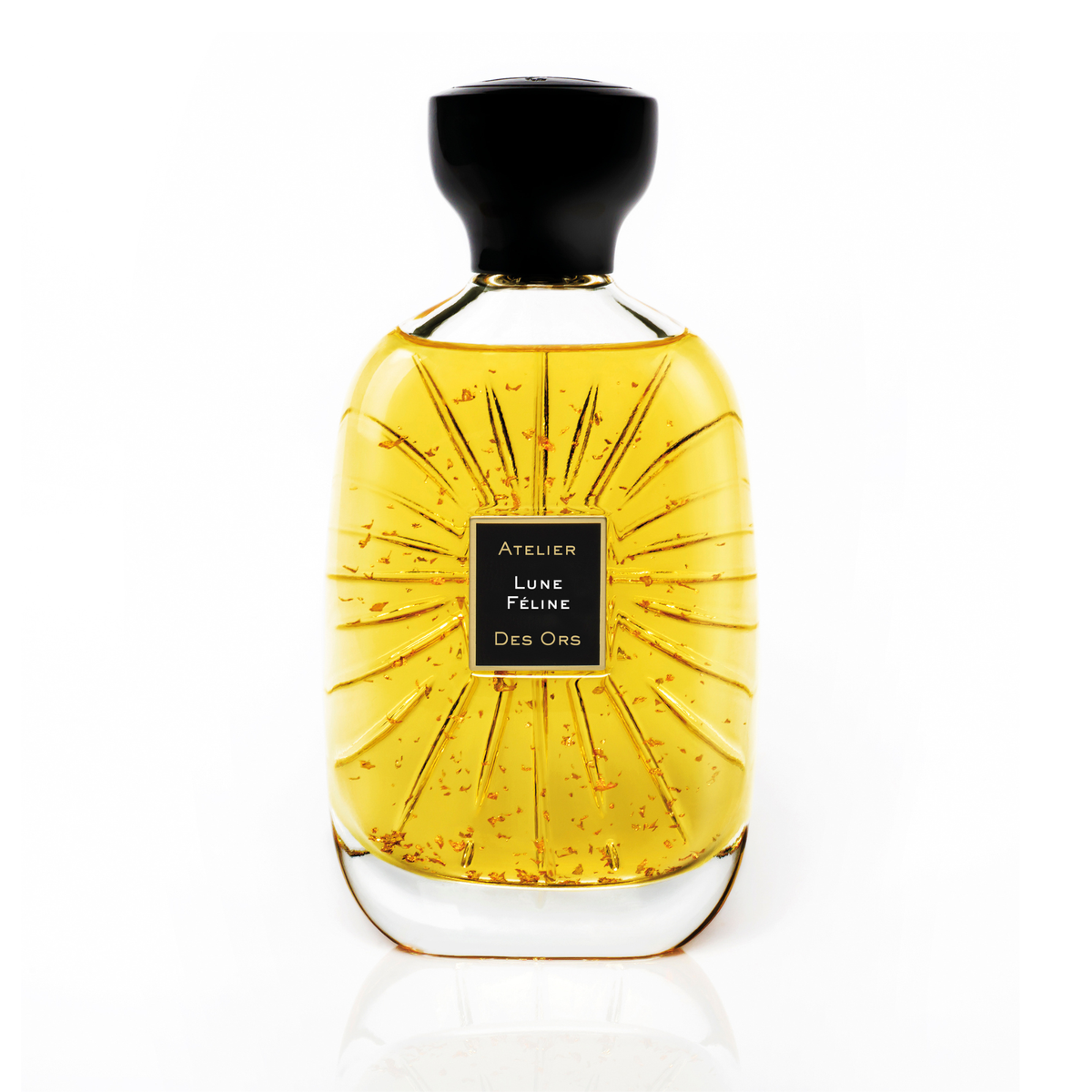 A clear glass perfume bottle filled with golden yellow liquid, featuring a black cap and a black central label with gold text reads Lune Feline from Atelier Des Ors. The bottle has a sunburst radial pattern on the front and is photographed on a clean white background with a subtle reflection below.