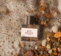 A rectangular glass perfume bottle labeled 'BOY BEACH BY ALEXANDER' with a black cap, lying on a bed of wet river stones and sea foam. The bottle is surrounded by white sea foam and multicolored pebbles.
