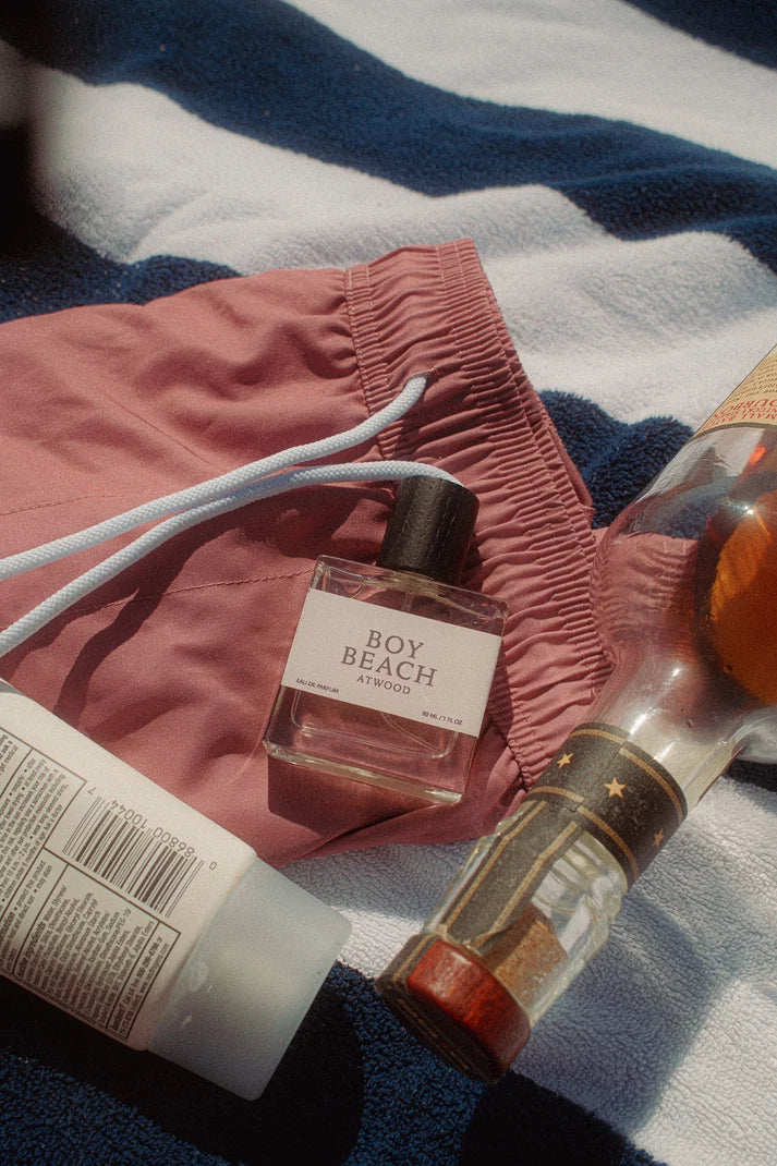 Perfume bottle labeled 'Boy Beach' on a towel with pink shorts and a bottle of lotion.