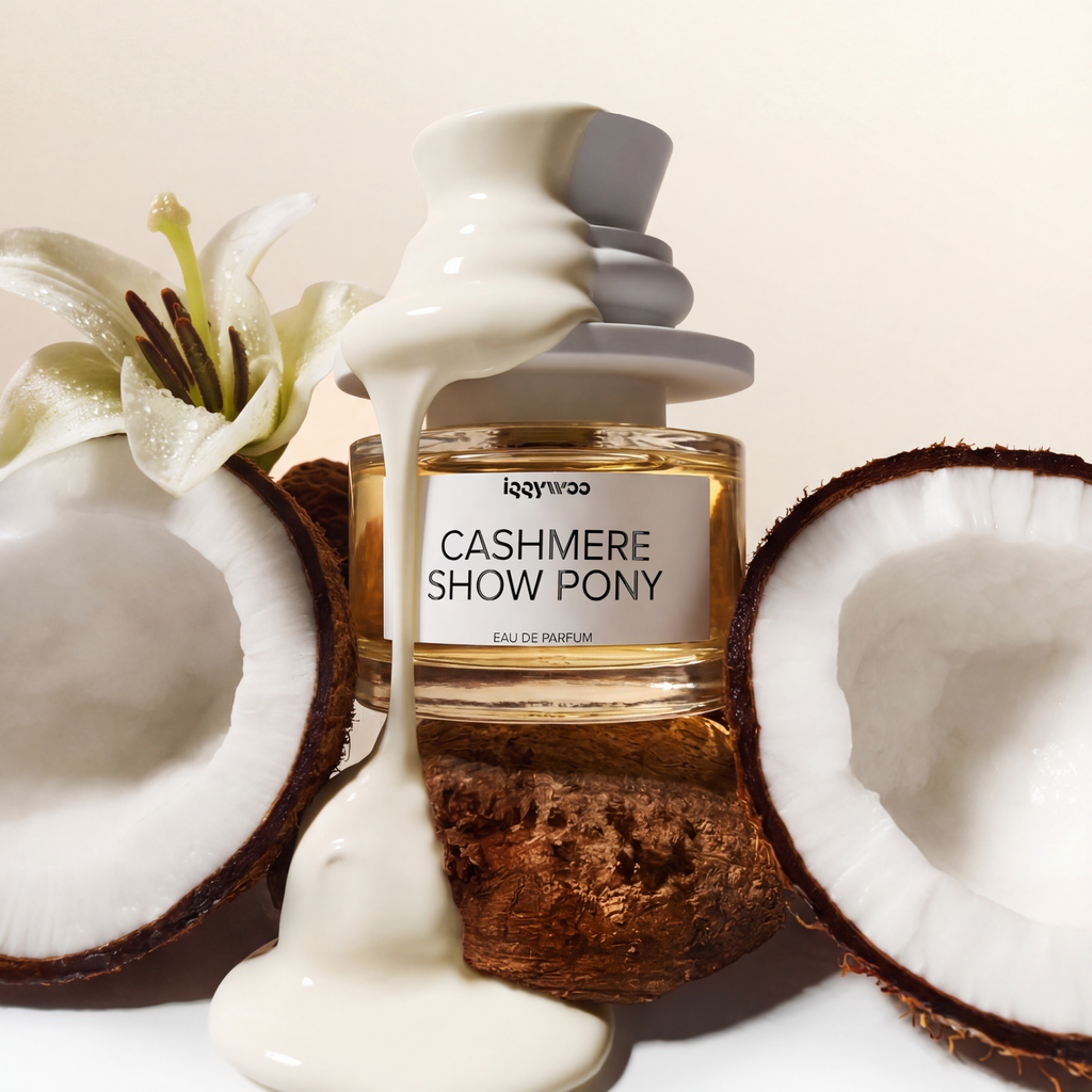 A perfume bottle labeled 'CASHMERE SHOW PONY' by the brand 'breezess', styled with white cream dripping from the cap, set between two halved coconuts and a white lily flower on a warm off-white background.