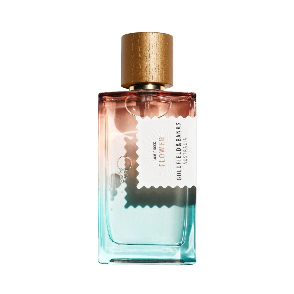 A rectangular glass perfume bottle with a gradient from pink at the top to light blue at the bottom, topped with a gold wood cap. A white label with black text reads Pacific Rock Flower from Goldfield and banks.