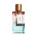 A rectangular glass perfume bottle with a gradient from pink at the top to light blue at the bottom, topped with a gold wood cap. A white label with black text reads Pacific Rock Flower from Goldfield and banks.