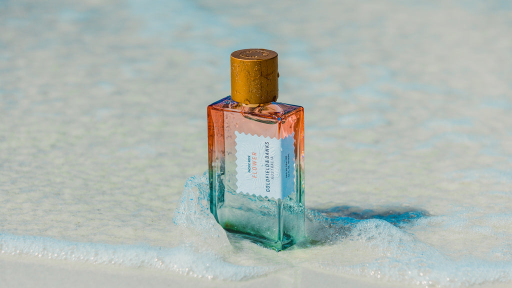 A rectangular glass perfume bottle with a gold wood cap, featuring a gradient of orange, pink, and teal blue, standing in shallow foamy water on a sandy beach.