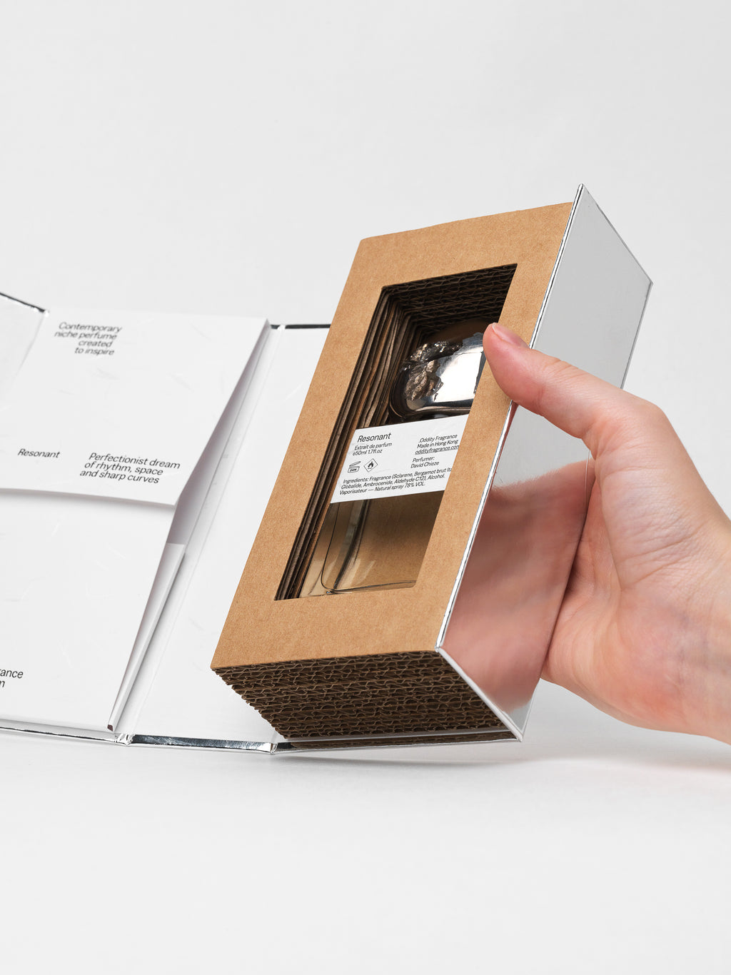 A hand holding a rectangular brown kraft cardboard display box with a clear window, showing a small round watch inside, against a clean white background with an open white booklet nearby.