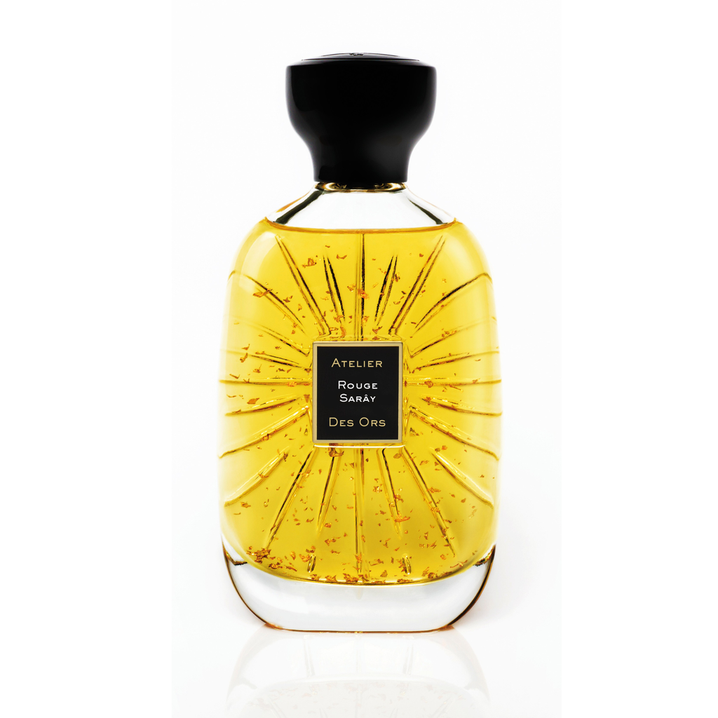 A clear glass perfume bottle filled with golden-yellow liquid, featuring a black cap and a central black label that reads 'ATELIER  DES ORS'ROUGE SARAY. The bottle has a textured, sunburst-like pattern on the front.